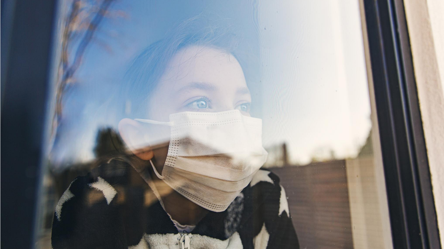Une enfant portant un masque derrière une vitre.