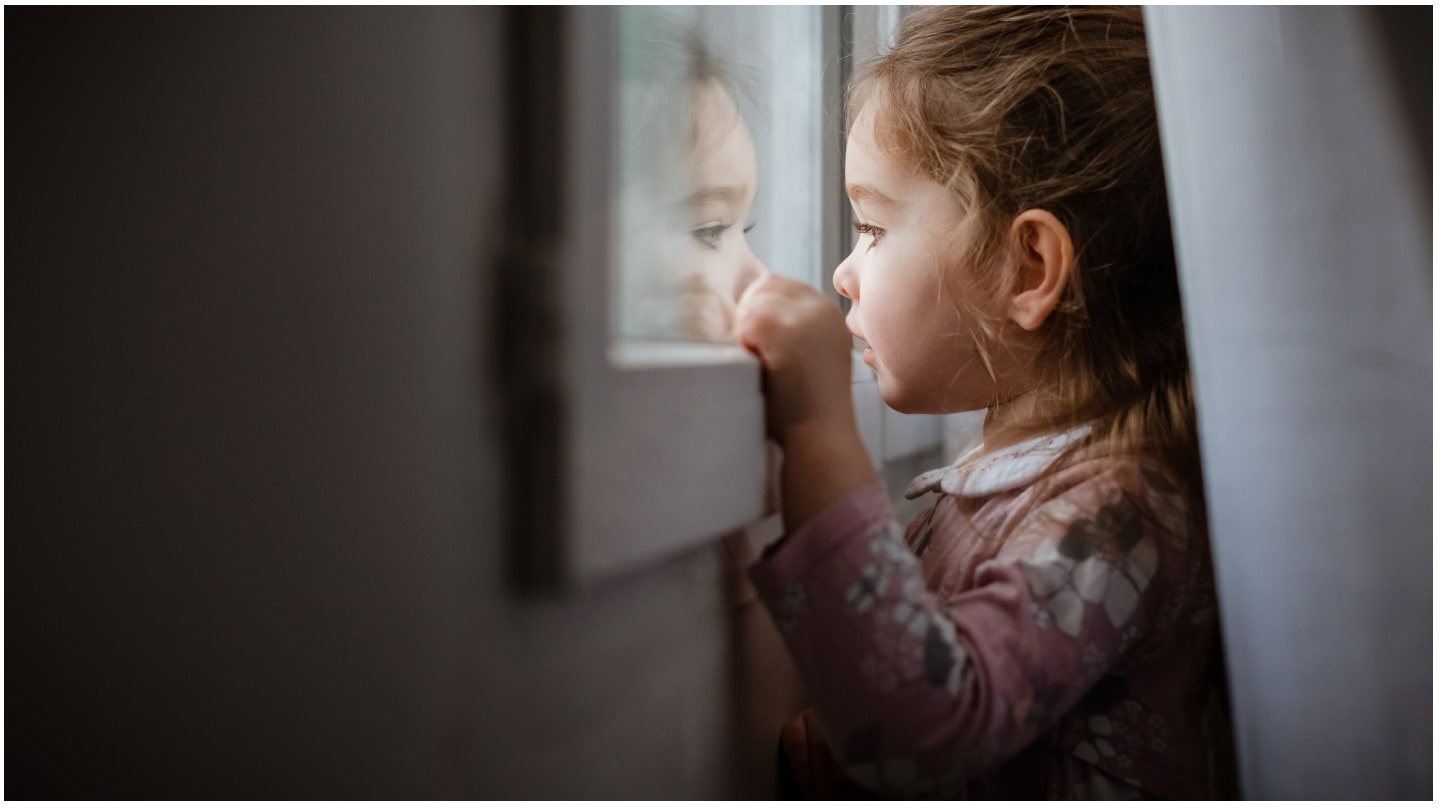 Une petite fille cachée derrière un rideau regarde par la fenêtre.