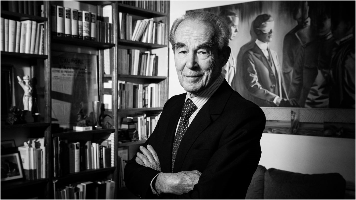 Robert Badinter souriant, les bras croisés, avec une bibliothèque en arrière-plan. Image en noir et blanc.