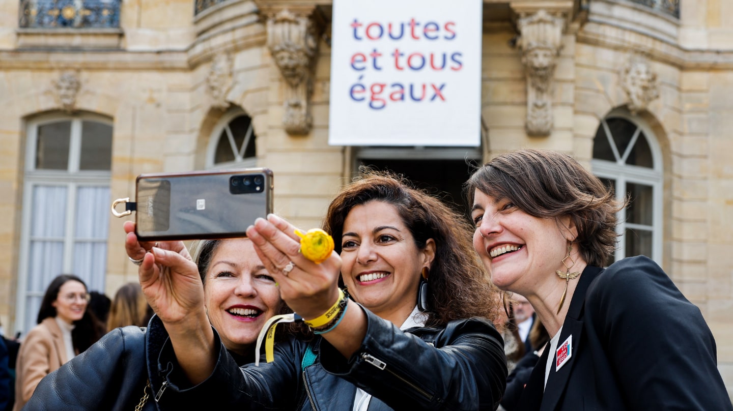 Trois femmes se prennent en photo dans la cours de l'Hôtel de Matignon. Une affiche "toutes et tous égaux" en fond sur la façade.